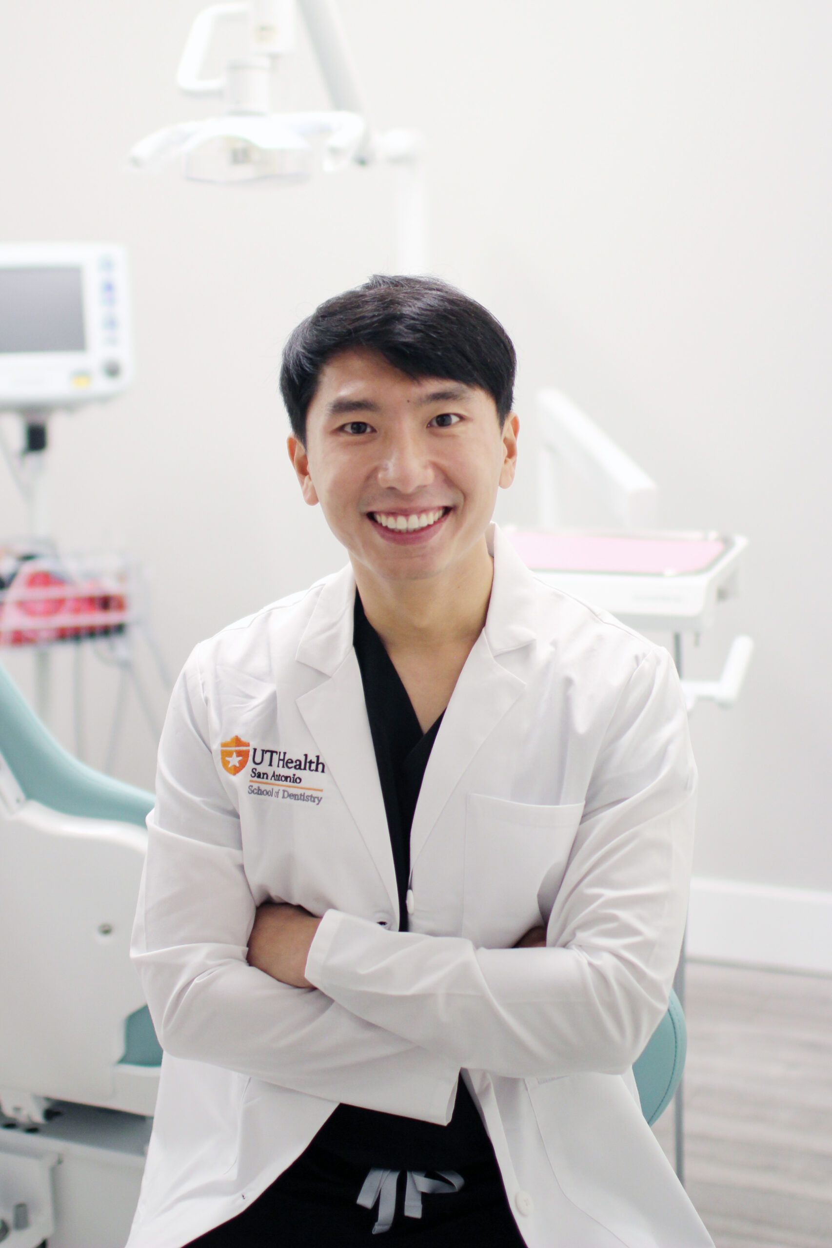 Dr. Marco Waworuntu, DDS, wearing a white coat with "UTHealth" branding, standing with arms crossed in a dental clinic setting, featuring dental equipment in the background - Puyallup Dentist Dr. Marco Waworuntu, DDS, wearing a white coat with "UTHealth" branding, standing with arms crossed in a dental clinic setting, featuring dental equipment in the background - Puyallup Dentist