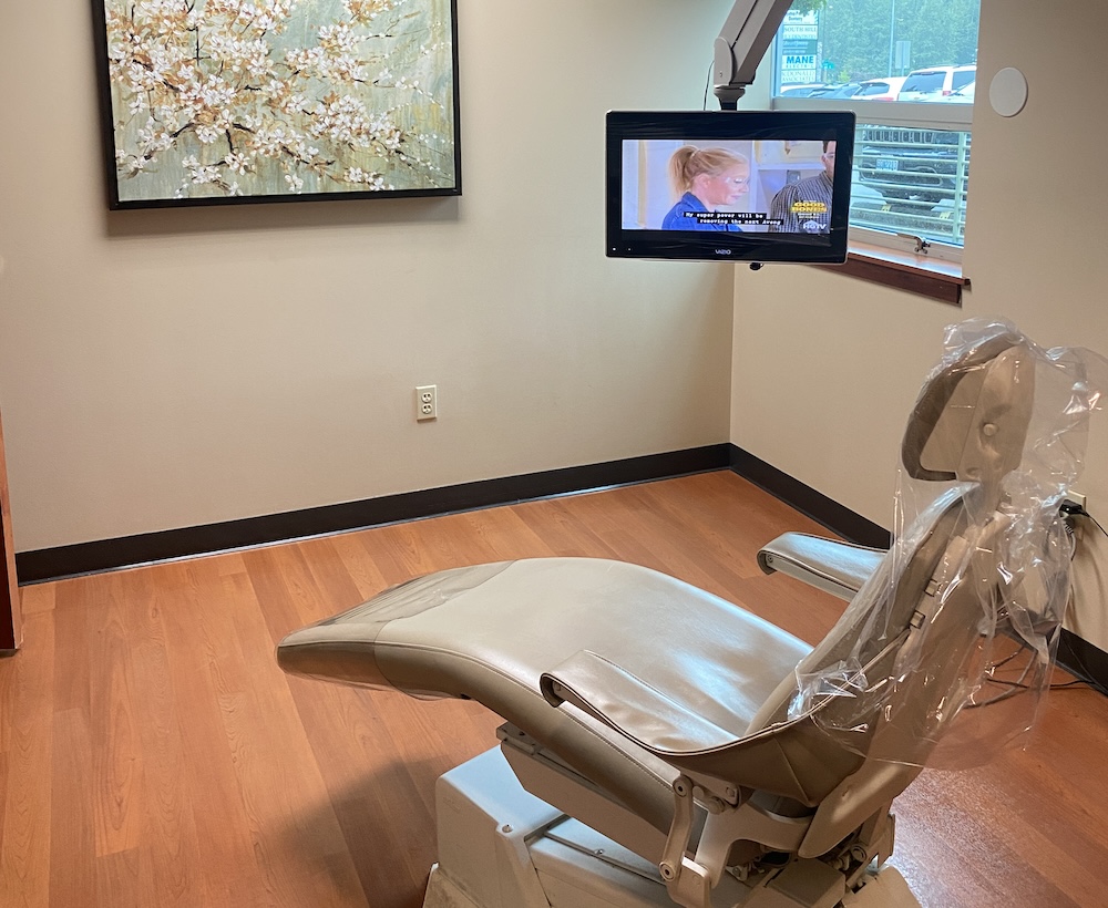 Dental operatory with a beige dental chair, wall-mounted TV showing a program, a window with natural light, and a decorative painting on the wall - Puyallup Dentist Dental operatory with a beige dental chair, wall-mounted TV showing a program, a window with natural light, and a decorative painting on the wall - Puyallup Dentist