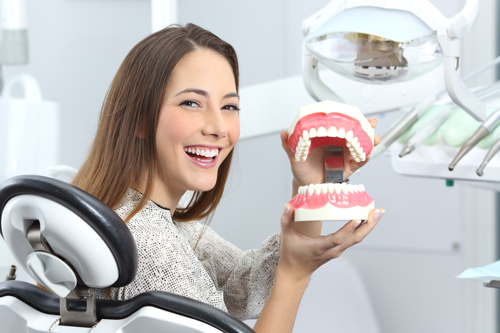 A smiling woman seated in a dental chair holding a dental model of teeth, with dental equipment visible in the background - Puyallup Dentist A smiling woman seated in a dental chair holding a dental model of teeth, with dental equipment visible in the background - Puyallup Dentist