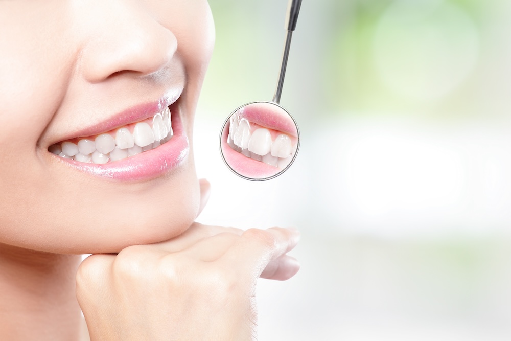 A close-up of a smiling woman's teeth with a dental mirror reflecting her teeth, set against a softly blurred green background - Puyallup Dentist A close-up of a smiling woman's teeth with a dental mirror reflecting her teeth, set against a softly blurred green background - Puyallup Dentist