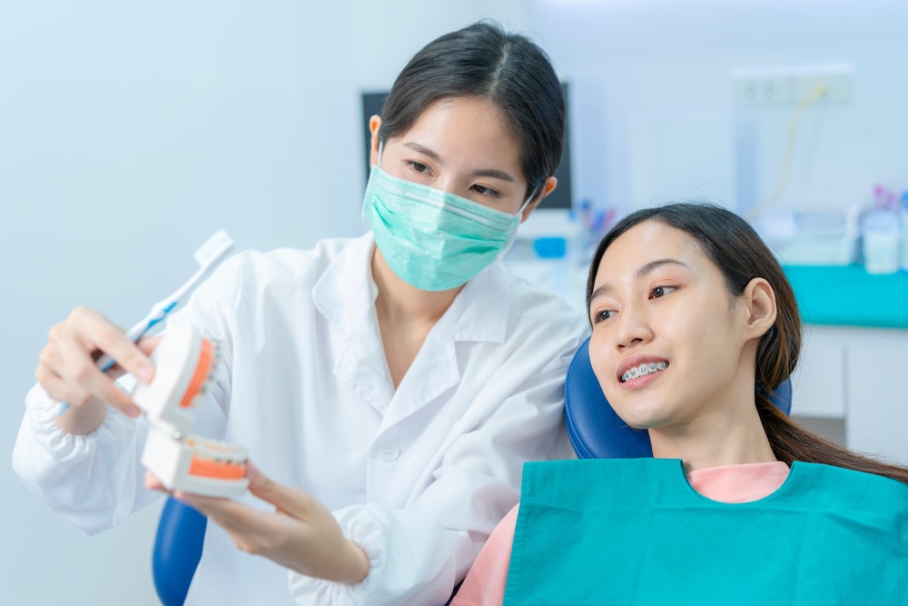 A dentist wearing a mask demonstrating proper brushing technique using a dental model to a patient with braces, seated in a dental chair - Puyallup Dentist A dentist wearing a mask demonstrating proper brushing technique using a dental model to a patient with braces, seated in a dental chair - Puyallup Dentist